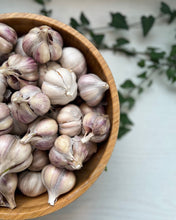 A wooden bowl filled with whole garlic bulbs, photographed from above on a light surface with blurred green leaves in the background.
